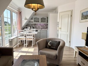 A modern living room with a sofa, a dining table with chairs, a kitchen area with cabinets, and a view of the outdoors through large windows. - Süderstraße, Apartment 12 – Landhaus - Cozy 3-room vacation rental for 4 (Sylt)