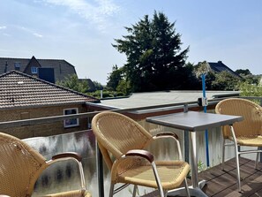 Eine Dachterrasse mit Korbstühlen und einem Tisch, mit Blick auf ein Wohngebiet mit Häusern und einem großen Baum.