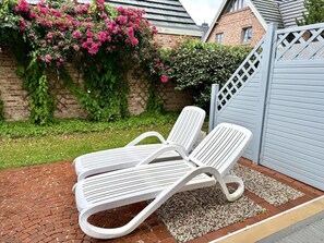 En terrasse med to hvide liggestole, en brobelagt sti og en mur med lyserøde blomster.