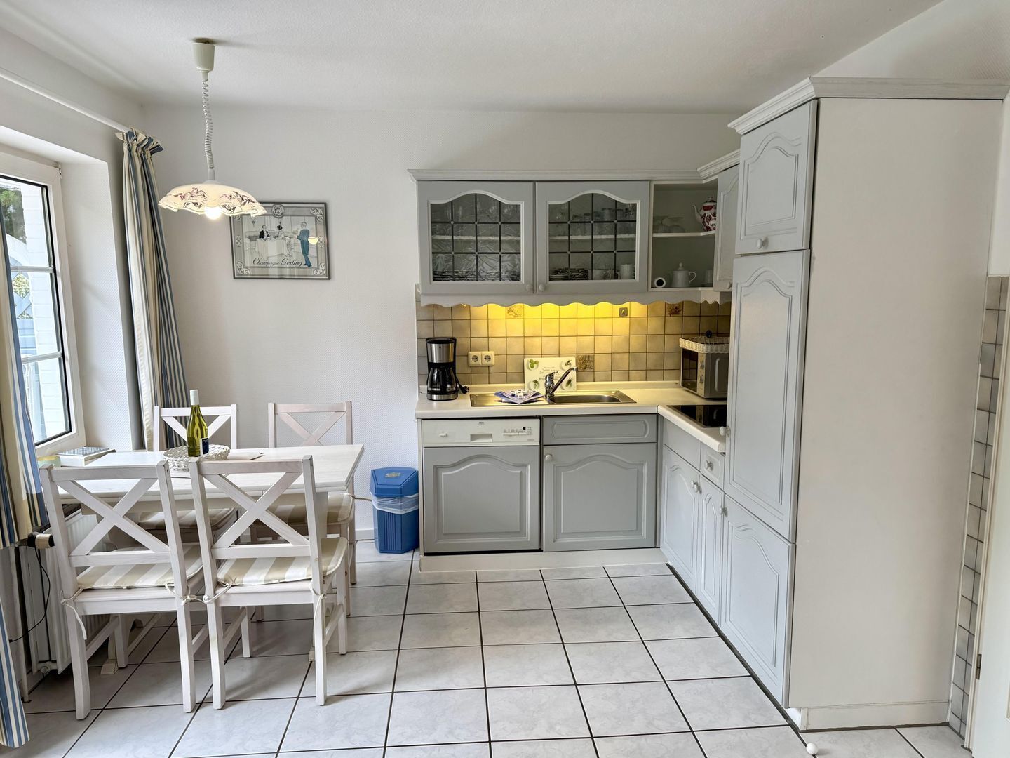 Una cocina con armarios blancos, una mesa de comedor con sillas, una cafetera y un cubo de basura.