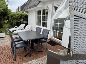 En udendørs terrasse med et bord og stole, et hvidt hus med et vindue og et overdækket grillområde.