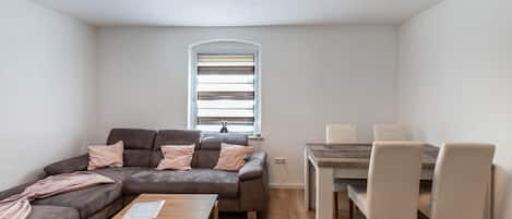A modern living room with a sectional sofa, a wooden coffee table, a dining table with chairs, and a window with blinds.