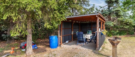 Ein überdachter Sitzbereich im Freien mit Tisch und Stühlen, umgeben von Bäumen und einem eingezäunten Garten.