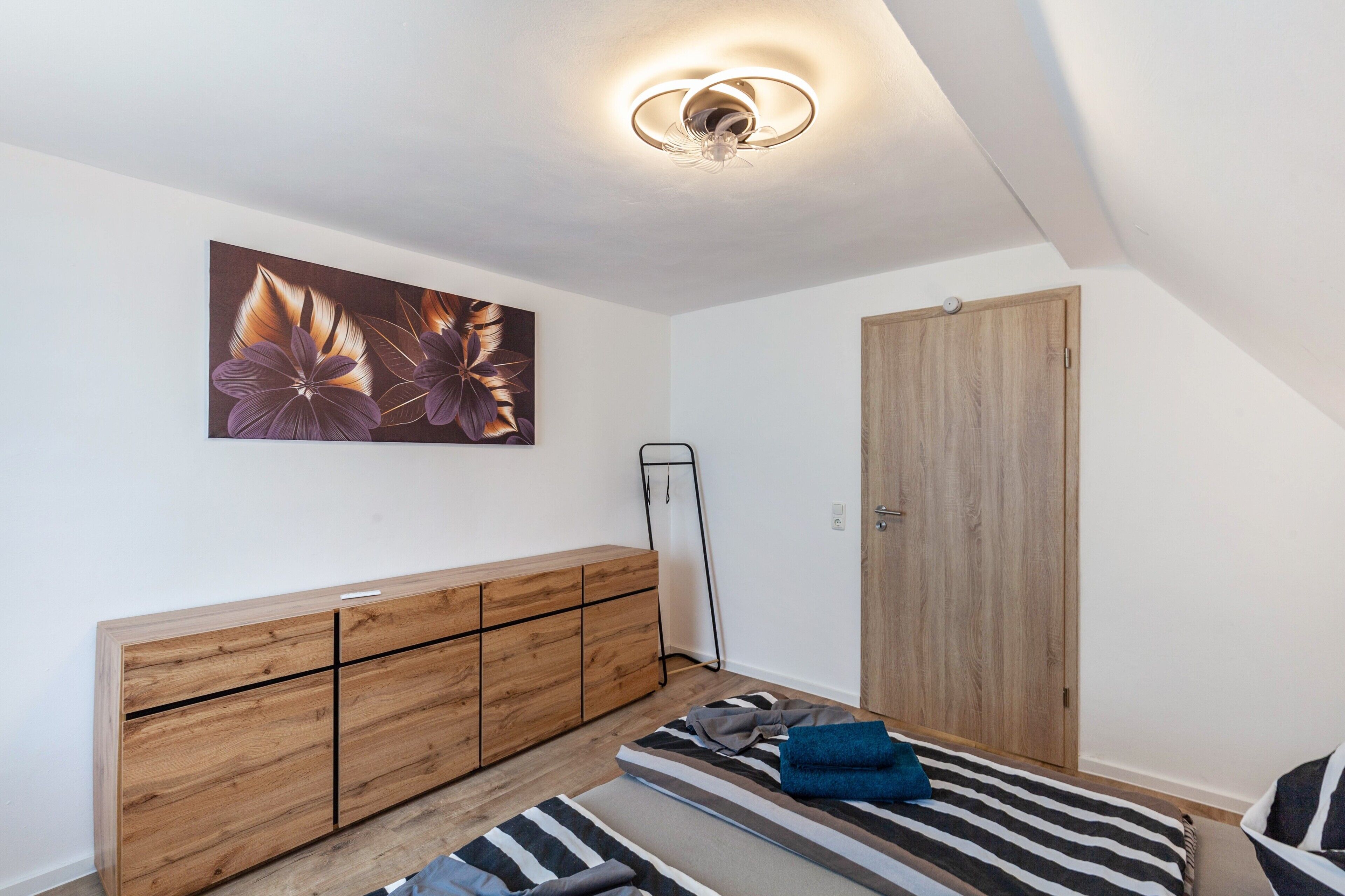Ein Schlafzimmer mit einem Holzkleiderschrank, einem Bett mit gestreifter Bettwäsche, einem Wandbild und einer Deckenleuchte.