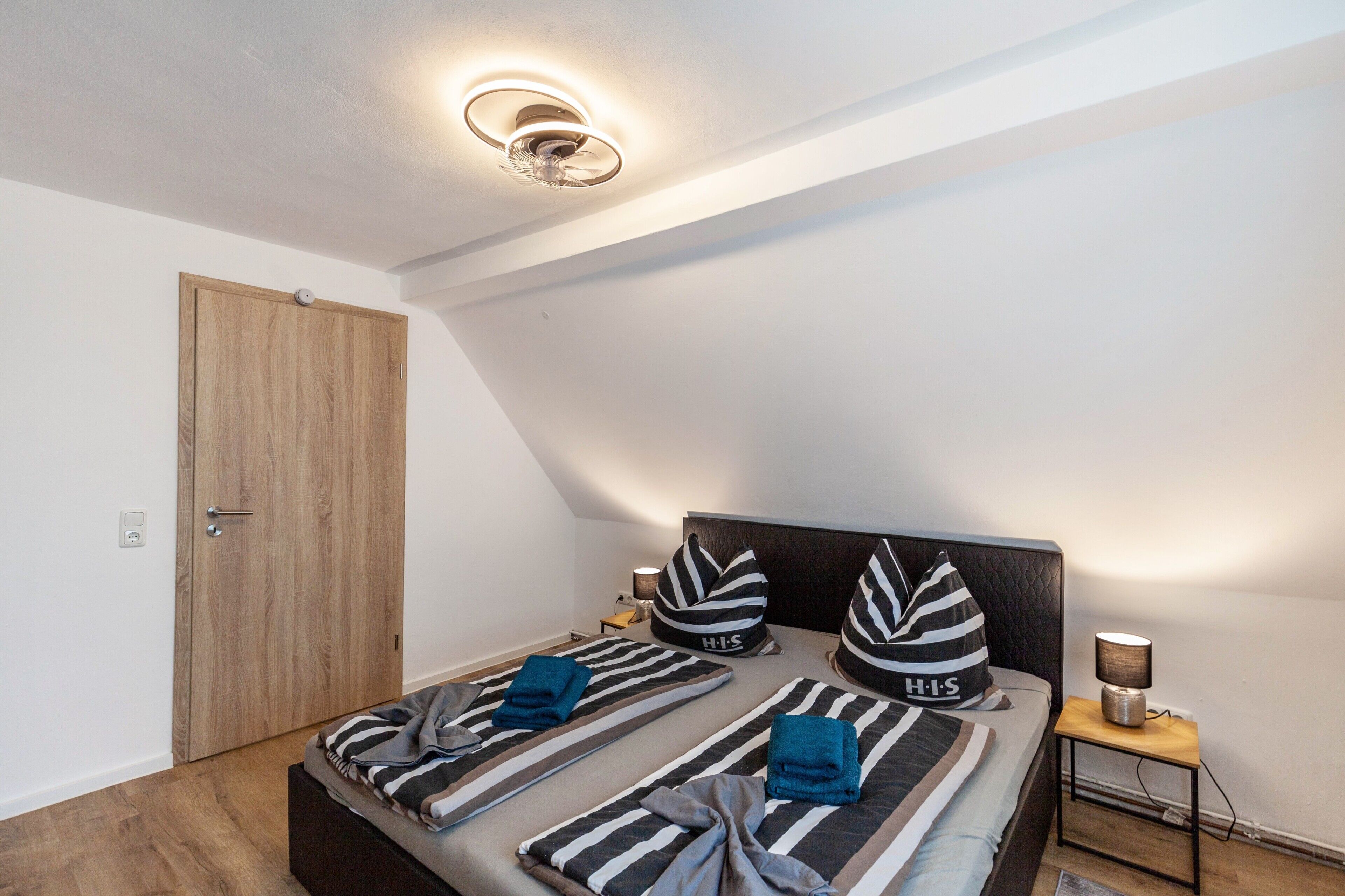 A bedroom with a bed, pillows, a bedside table, a lamp, and a wooden door.