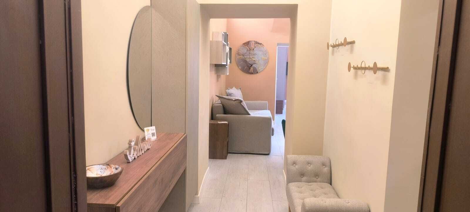 Un corridoio che conduce a una zona giorno con un divano e una poltrona, un tavolo consolle in legno con un cartello di benvenuto e una sveglia appesa al muro.