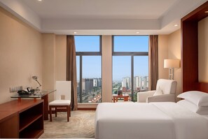 Habitación de hotel con una cama grande, dos sillones, un escritorio con lámpara y vistas a la ciudad a través de amplias ventanas.