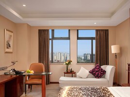 Habitación de hotel con escritorio, silla, sofá y vistas a la ciudad a través de amplios ventanales.