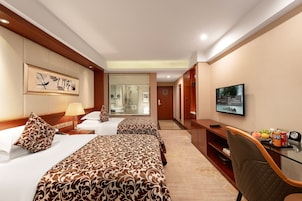 Habitación de hotel con dos camas, televisión, escritorio con silla y baño visible a través de una puerta de vidrio.