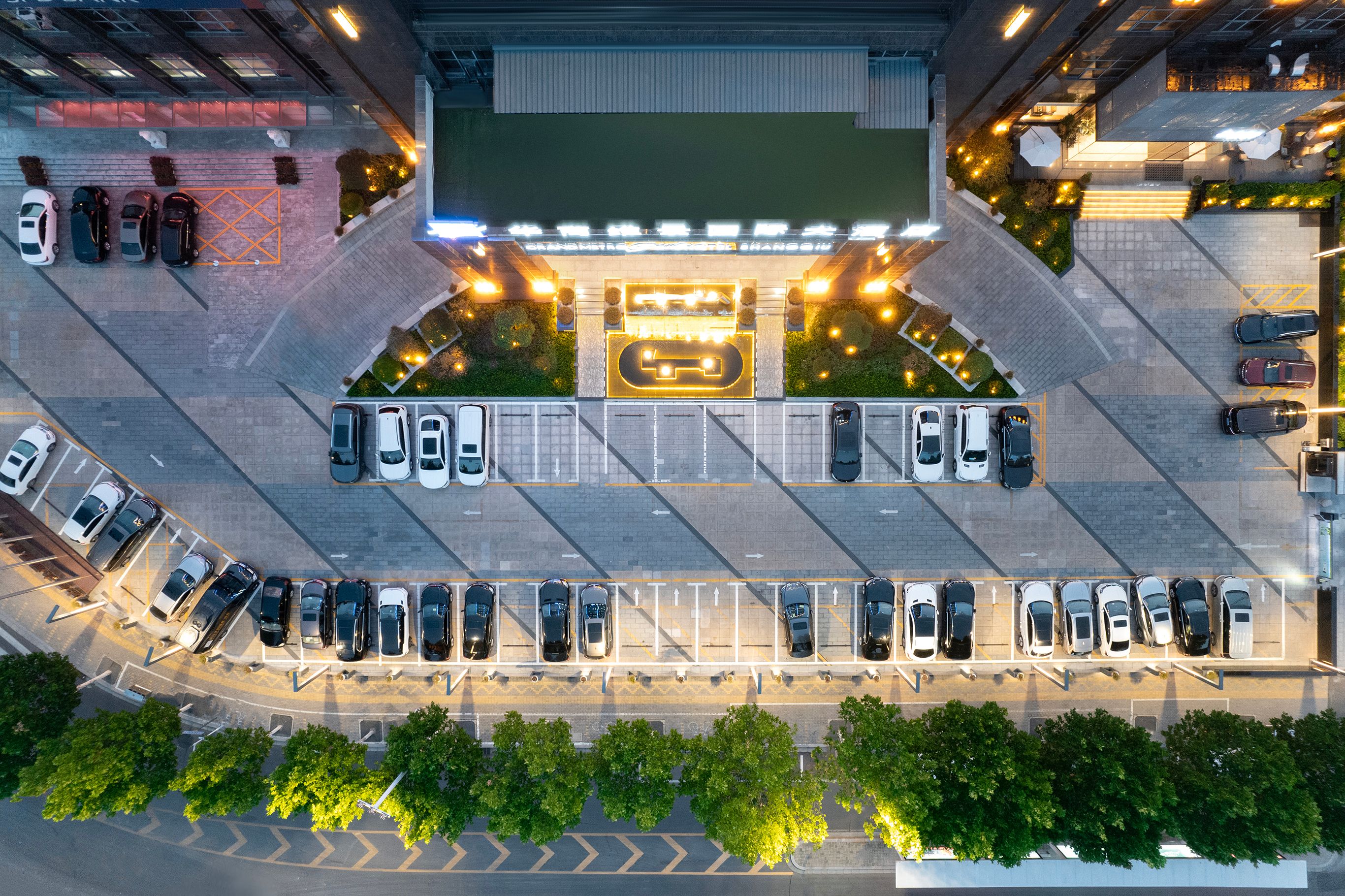 Vista aérea de un estacionamiento con varias filas de automóviles, un edificio con señalización iluminada y zonas verdes circundantes.