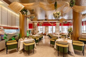 A restaurant with multiple tables, green upholstered chairs, and a chef statue.