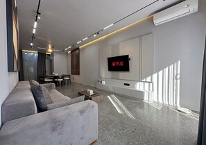 A modern living room with a grey sofa, a flat-screen TV mounted on the wall, and a dining area with a table and chairs. - Modern 2BR Villa Umalas Great Location (Umalas)