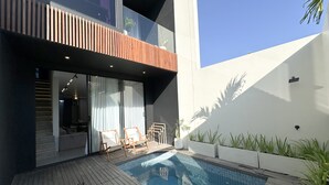 Un área de piscina exterior moderna con un deck de madera, dos sillas de descanso y una puerta de vidrio que da a un espacio interior.