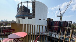 Blick auf eine Baustelle mit einer großen weißen, zylindrischen Struktur, einem Kran und einem Gewässer mit Booten. Auf einem Balkon stehen ein roter Tisch und Stühle.