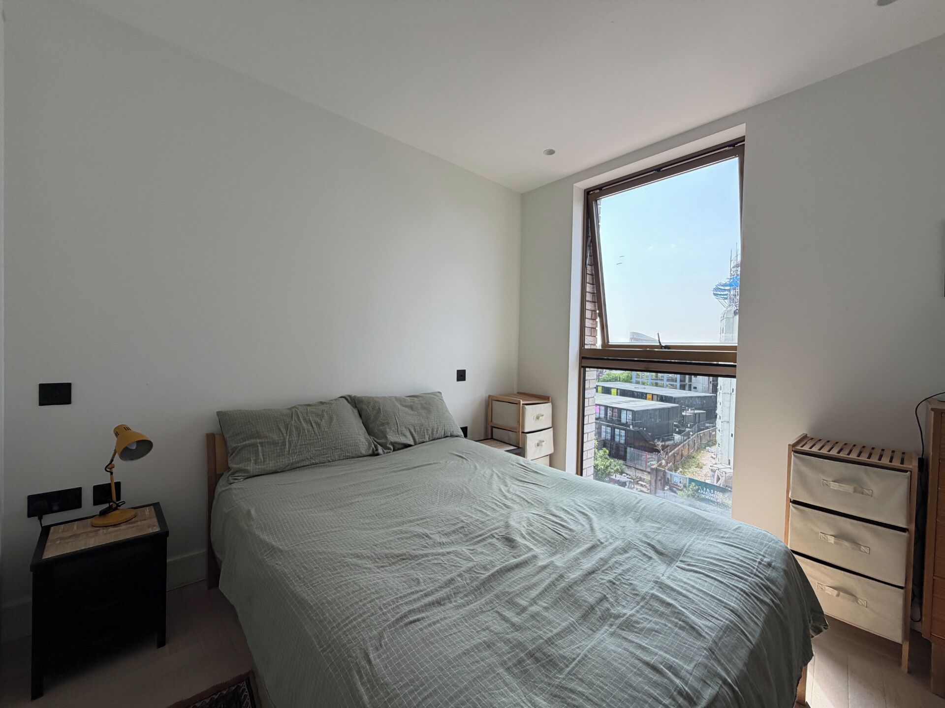 Une chambre avec une grande fenêtre, un lit, une table de chevet, une lampe et une commode.