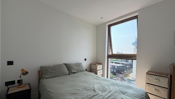 Ein Schlafzimmer mit großem Fenster, einem Bett, einem Nachttisch, einer Lampe und einer Kommode.