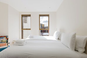 A hotel room with two beds, a view of the city through large windows, and a bookshelf with books. - Stylish Canal Side 2BDR Flat with Private Balcony (Greater London)