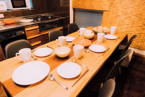 Mesa de comedor con platos, tazones, tazas y cubiertos, rodeada de sillas en una cocina moderna con electrodomésticos.