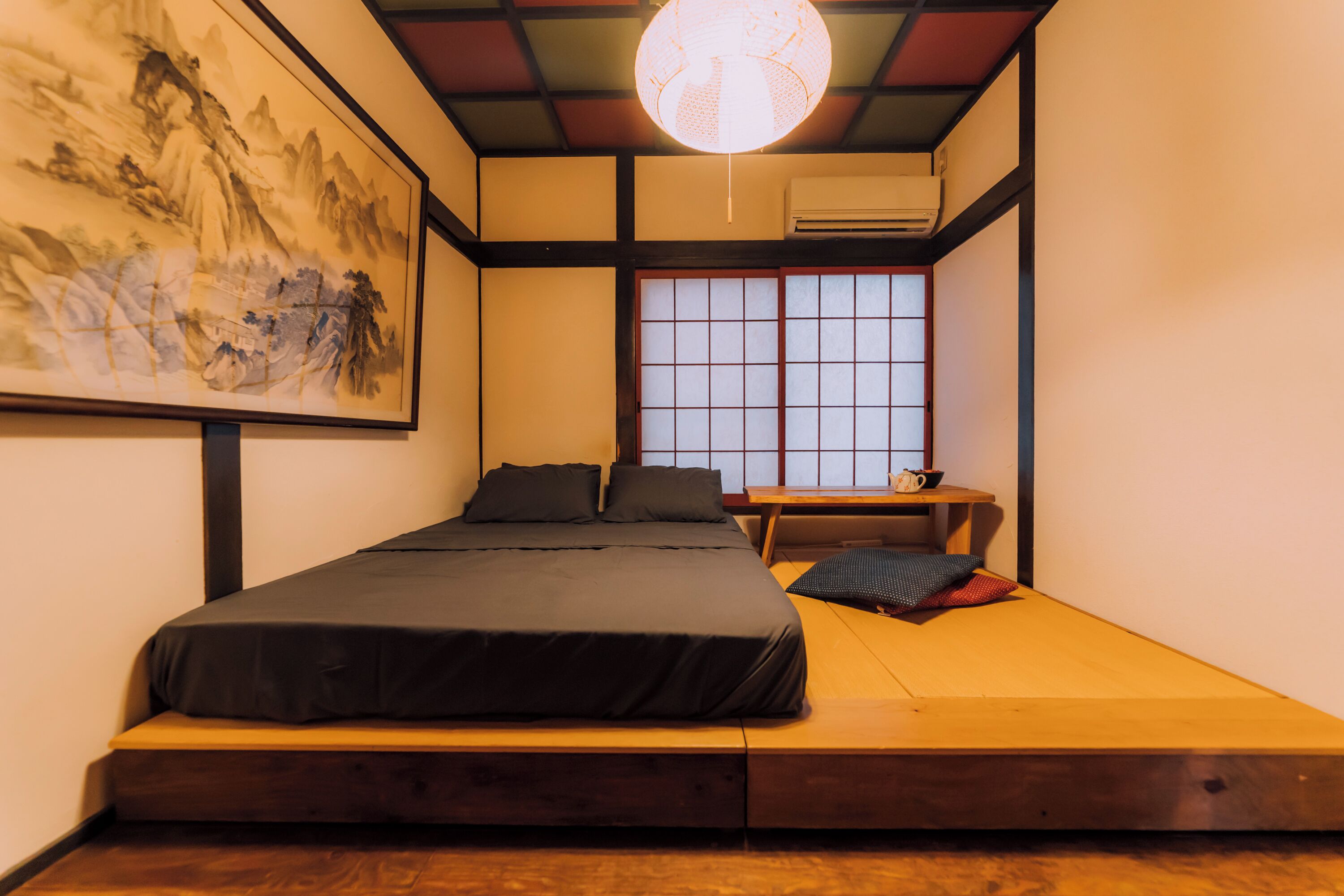 A room with a bed, a small table, a window with a view, and a traditional painting on the wall.