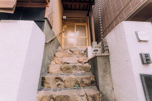 A stone staircase leading to a wooden door with a traditional design, a small outdoor area with a stone bench, and a wall-mounted light fixture. - Ninja 4BR Tennoji 5 Min (Osaka)