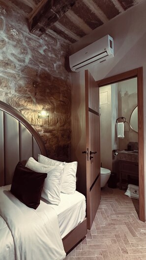 Un dormitorio con pared de piedra, una cama con ropa de cama blanca, una puerta de madera y un equipo de aire acondicionado.