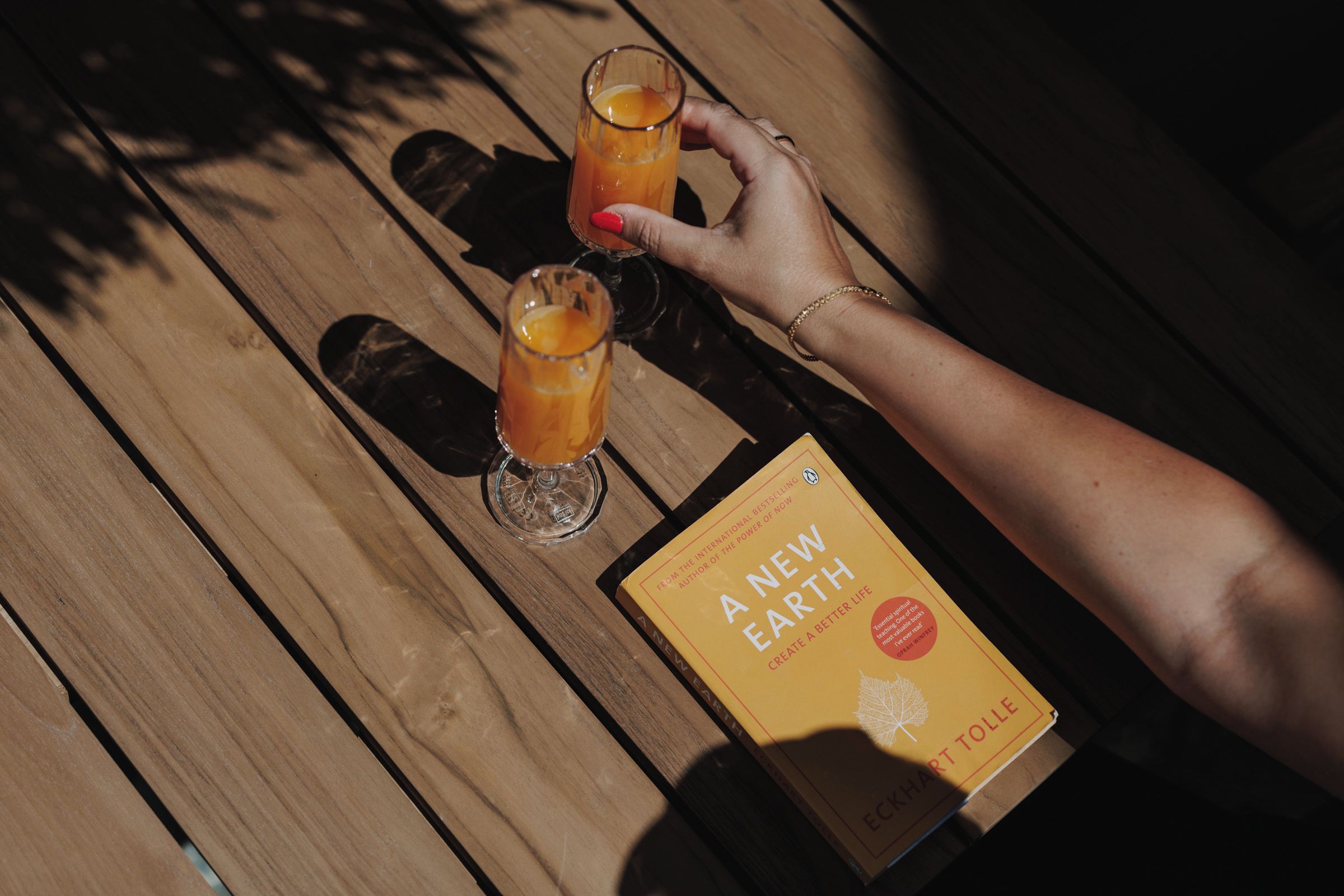 Une table en bois sur laquelle sont posés deux verres contenant une boisson à l’orange et un livre intitulé « Une nouvelle Terre » d’Eckhart Tolle.
