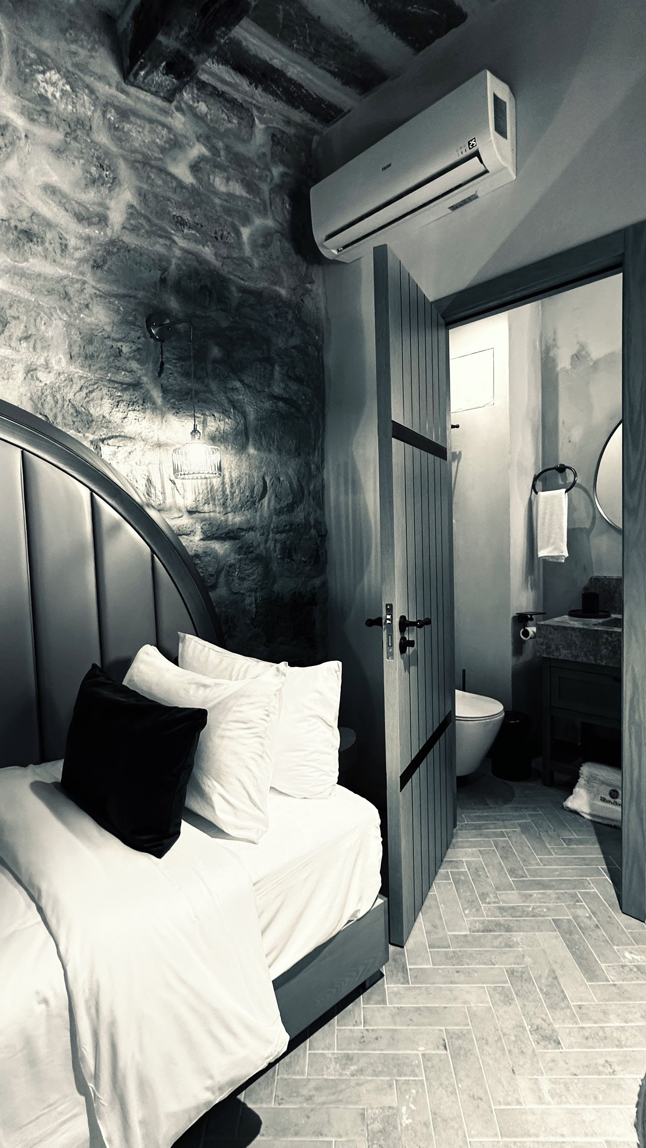 A bedroom with a stone wall, a bed with white bedding, an air conditioning unit, and a bathroom with a toilet and sink.