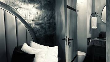 A bedroom with a stone wall, a bed with white bedding, an air conditioning unit, and a bathroom with a toilet and sink.
