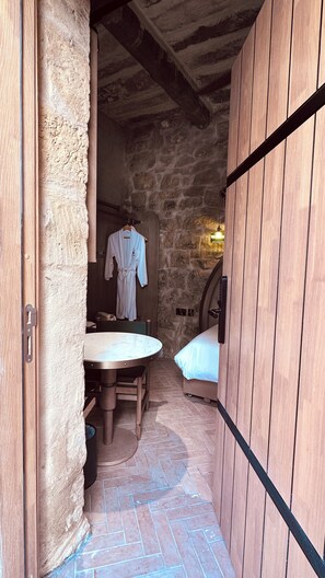 Una habitación pequeña de paredes de piedra con una cama, una mesa y una bata colgada en la pared.