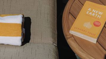 Un libro titulado 'Una Nueva Tierra' de Eckhart Tolle sobre una mesita de madera junto a un sofá con una toalla doblada.