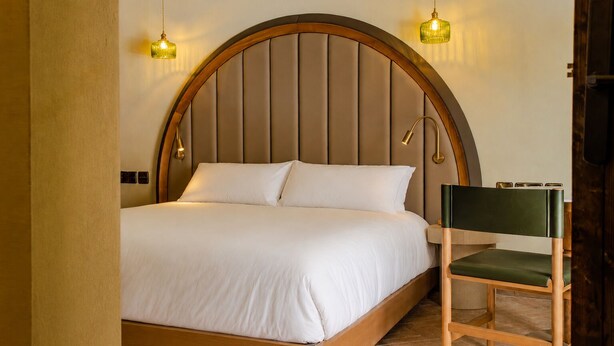 Una cama con estructura y cabecera de madera, dos lámparas de noche y una silla con cojín verde.