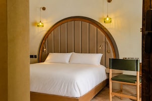 Uma cama com estrutura e cabeceira de madeira, duas luminárias de cabeceira e uma cadeira com almofada verde.