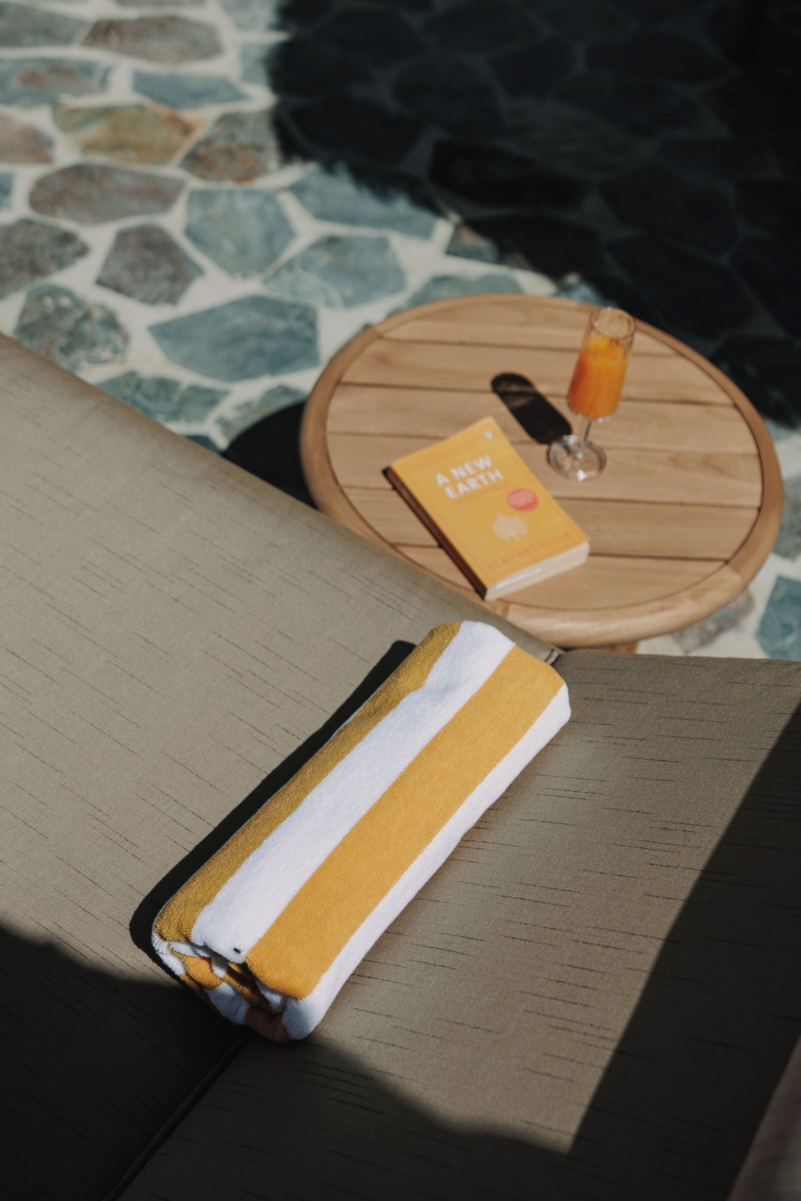 A sun lounger with a towel, a book titled 'A New Earth,' and a glass of orange juice on a round wooden table.