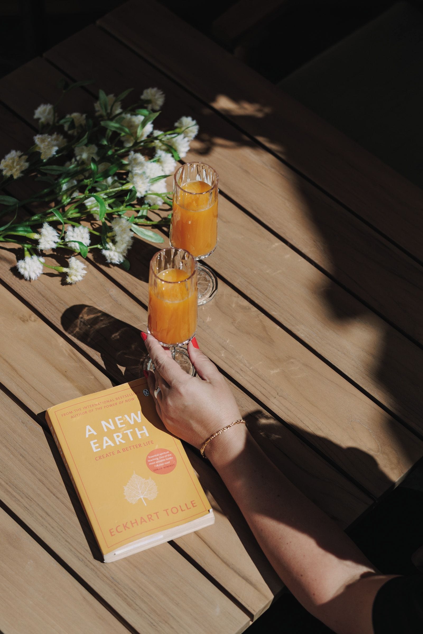 Un libro titulado 'A New Earth' de Eckhart Tolle sobre una mesa de madera con dos vasos de jugo de naranja.