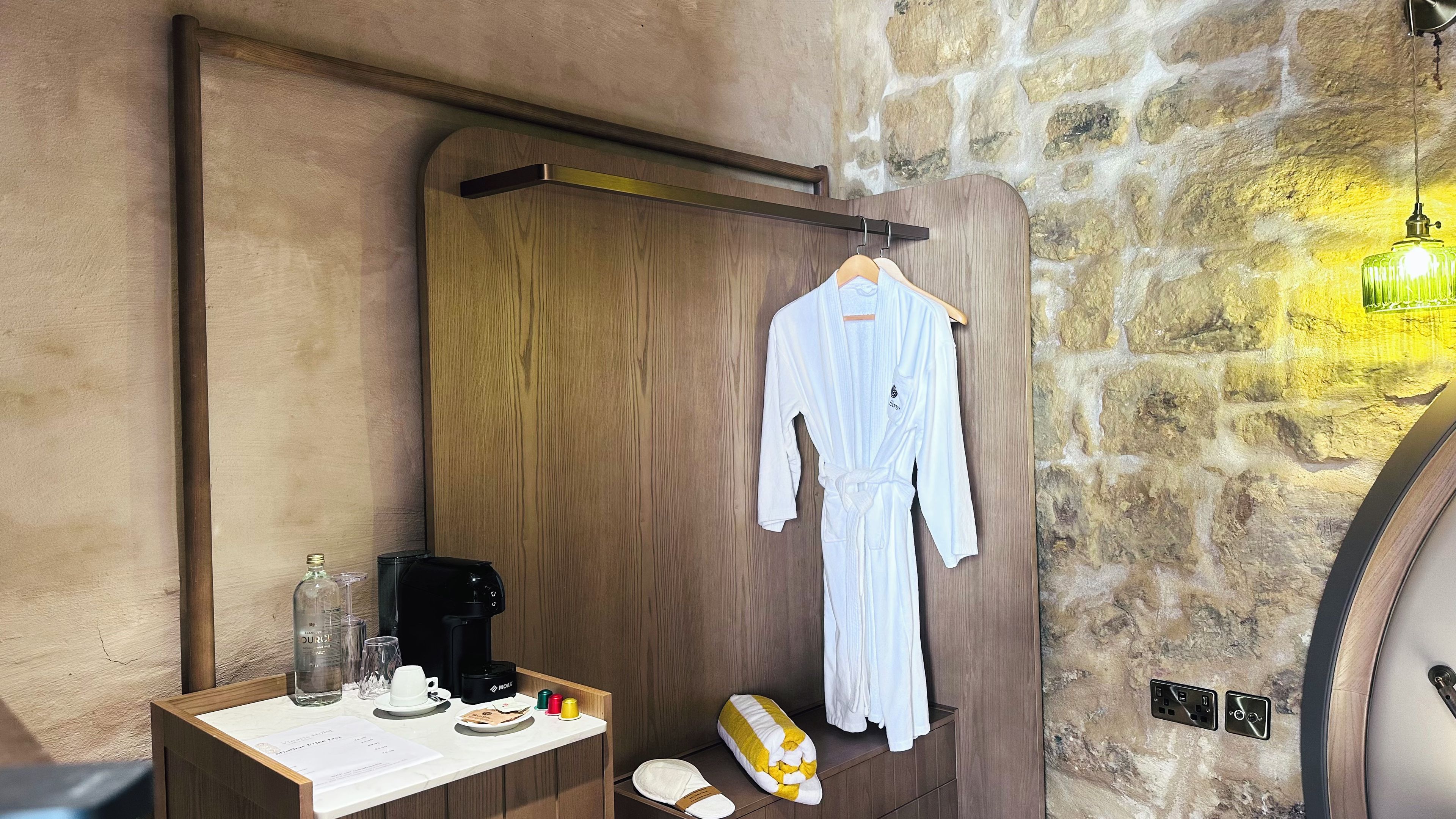 A bathroom with a wooden door, a robe hanging on a rack, a coffee maker, toiletries, and a stone wall.