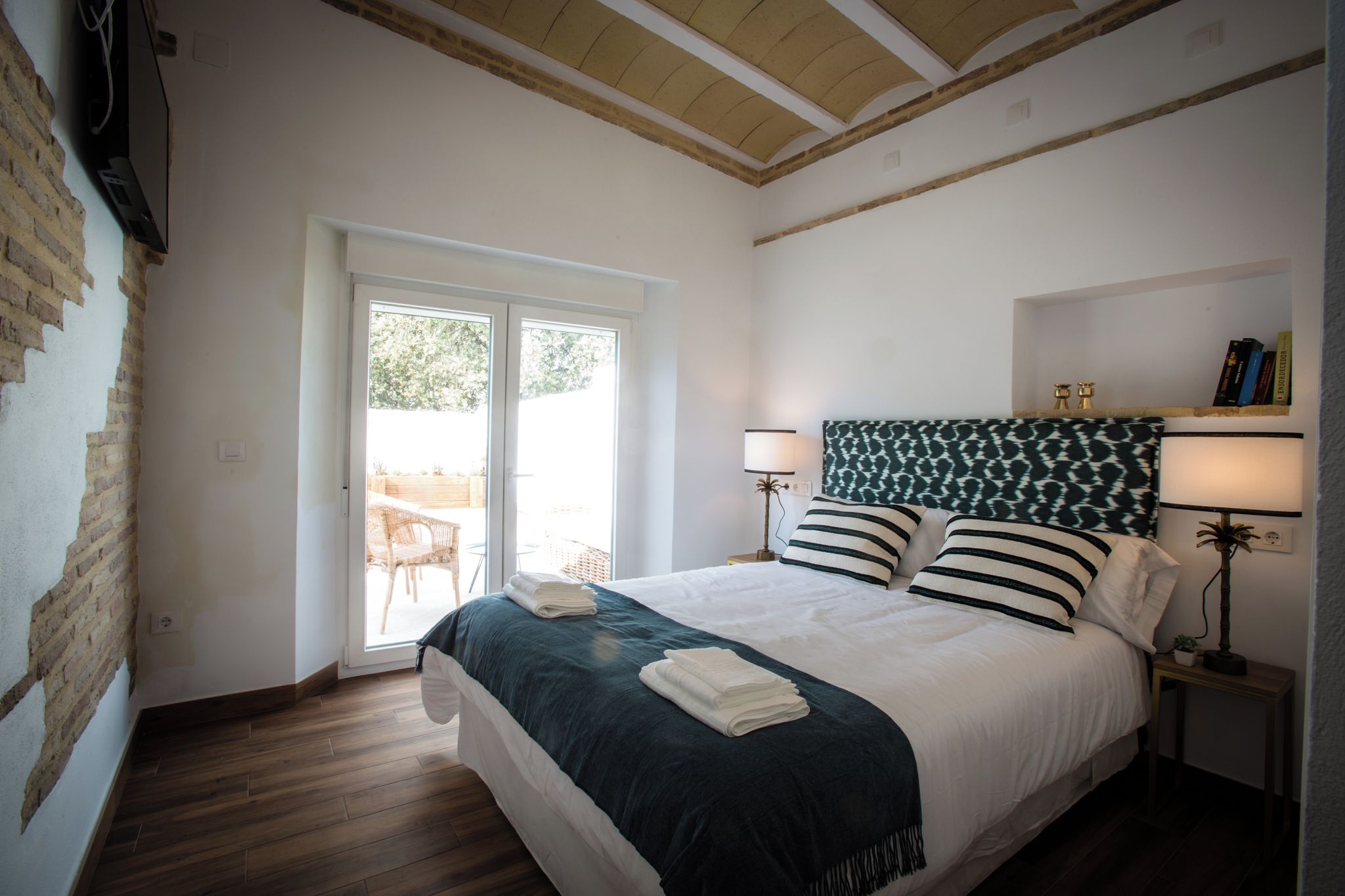 A hotel room with a bed, two bedside lamps, a bookshelf, and a view through a sliding door to an outdoor area.