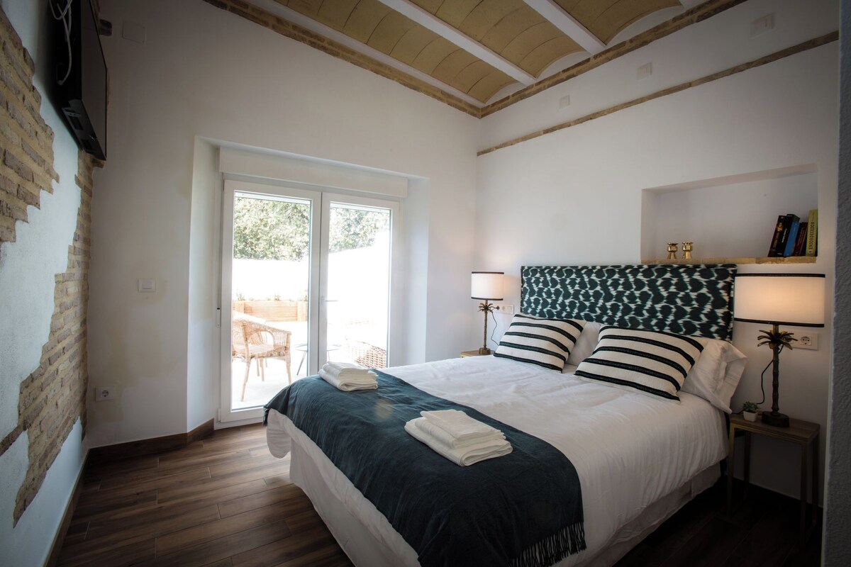 A hotel room with a bed, two bedside lamps, a bookshelf, and a view through a sliding door to an outdoor area.