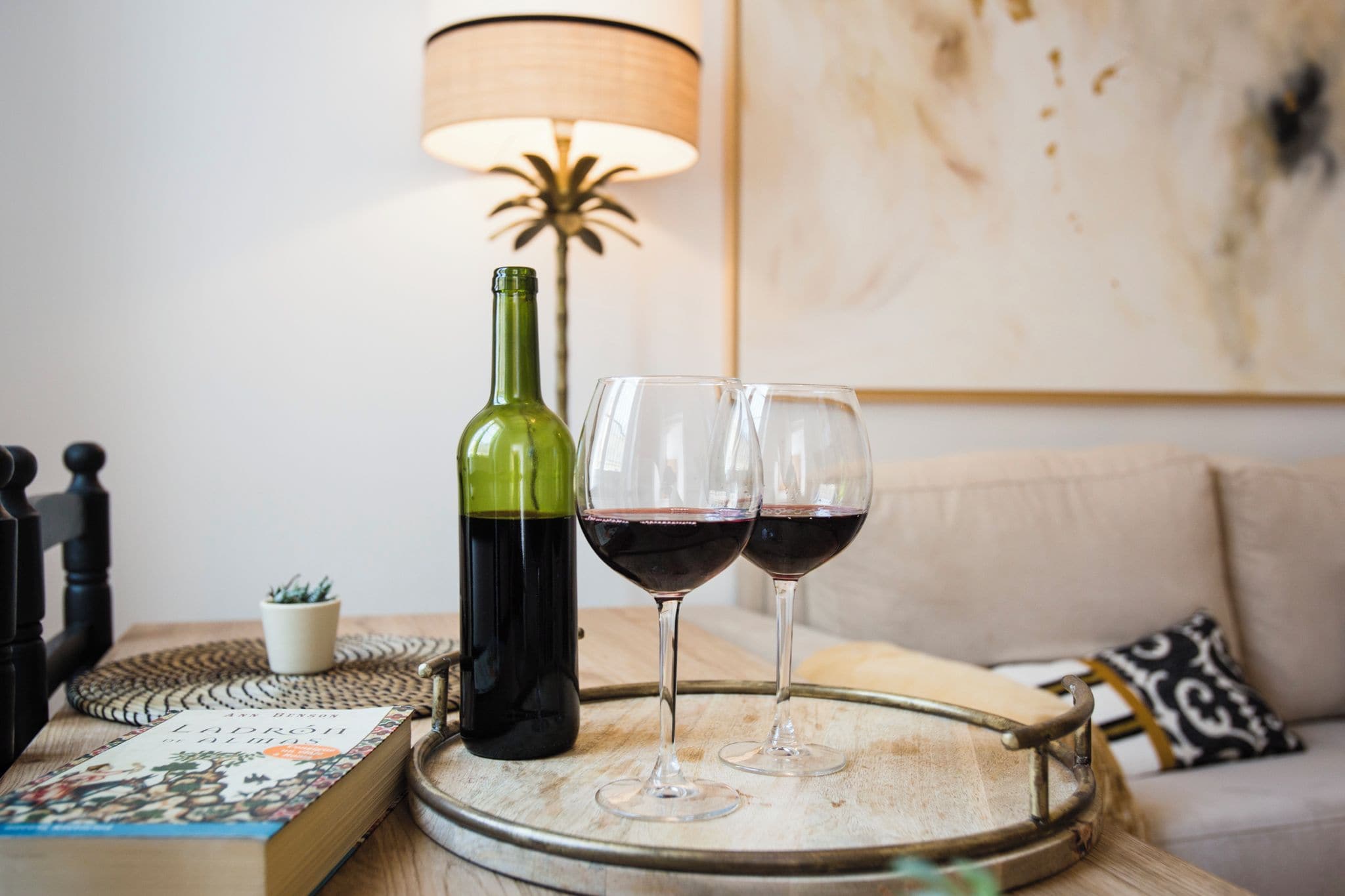 Une bouteille de vin, deux verres à vin, un plateau, un livre, une lampe et un canapé.