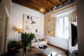 A modern living room with a large abstract painting, a wooden ceiling, a brick wall, a sofa, a coffee table, and a TV. - Alijar Casa Boutique (Castilleja de la Cuesta)