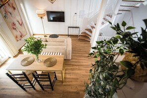 A modern living room with a sofa, a dining table with chairs, a TV mounted on the wall, and a staircase with a railing. - Alijar Casa Boutique (Castilleja de la Cuesta)