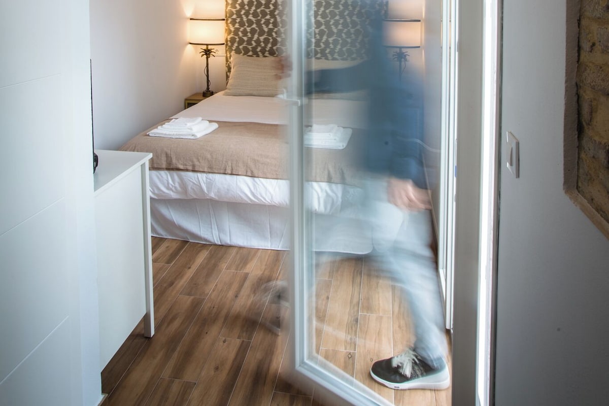 A bedroom with a bed, bedside table, lamp, and a person walking through a doorway.