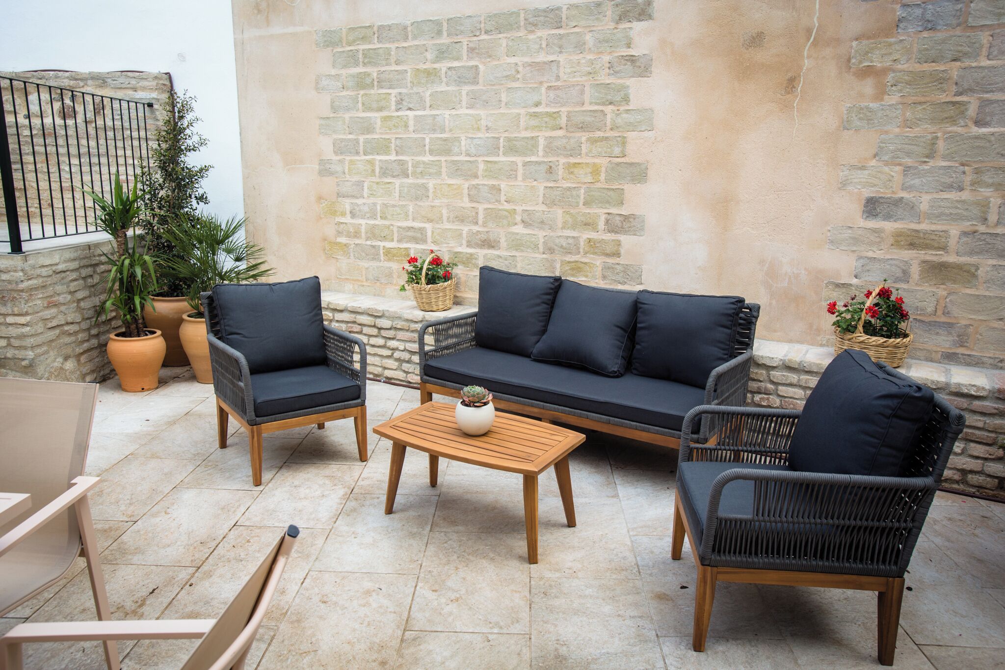 Un espacio al aire libre con mobiliario de mimbre, incluyendo dos sillones, un sofá y una mesa de centro. Hay plantas en macetas y una cesta con flores.