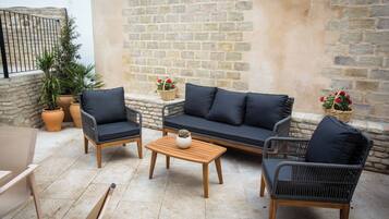 Un espacio al aire libre con mobiliario de mimbre, incluyendo dos sillones, un sofá y una mesa de centro. Hay plantas en macetas y una cesta con flores.