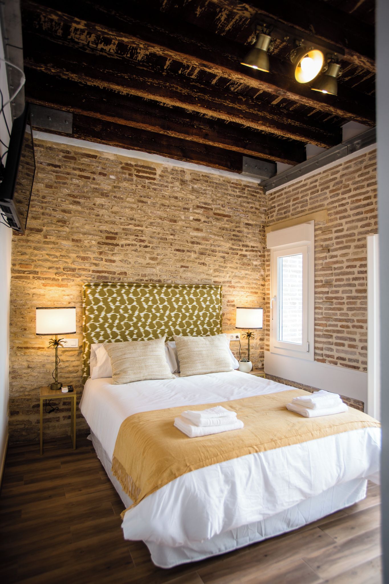 A hotel room with a brick wall, a bed with white linens, a green patterned headboard, and two bedside lamps.