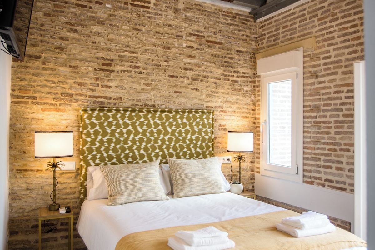 A hotel room with a brick wall, a bed with white linens, a green patterned headboard, and two bedside lamps.