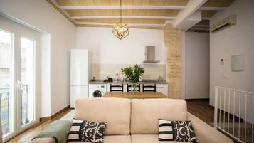 Una sala de estar con un sofá color beige, cojines estampados, una mesa de comedor y una cocina al fondo.