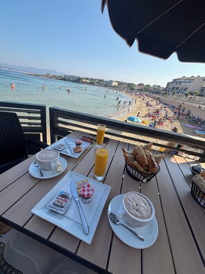 Ein Tisch mit Frühstücksangeboten und Blick auf einen Strand, wo Menschen schwimmen und sonnenbaden.