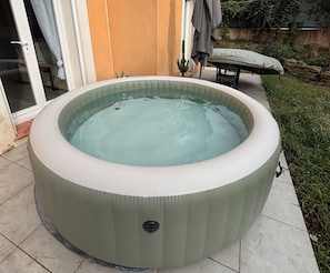 Una bañera de hidromasaje redonda con panel de control, ubicada en un patio con baldosas, zona verde y una pared de piedra al fondo.