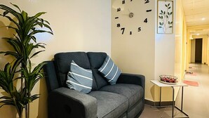 Una sala de estar con un sofá, una planta en maceta, un reloj de pared y una pequeña mesa con un frutero.