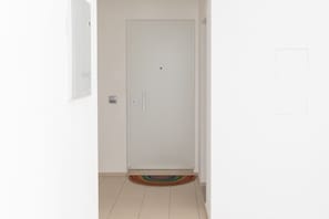 Eine weiße Tür mit Griff, links daneben ein kleines, rechteckiges Objekt an der Wand und ein farbenfroher, runder Teppich auf dem Boden.